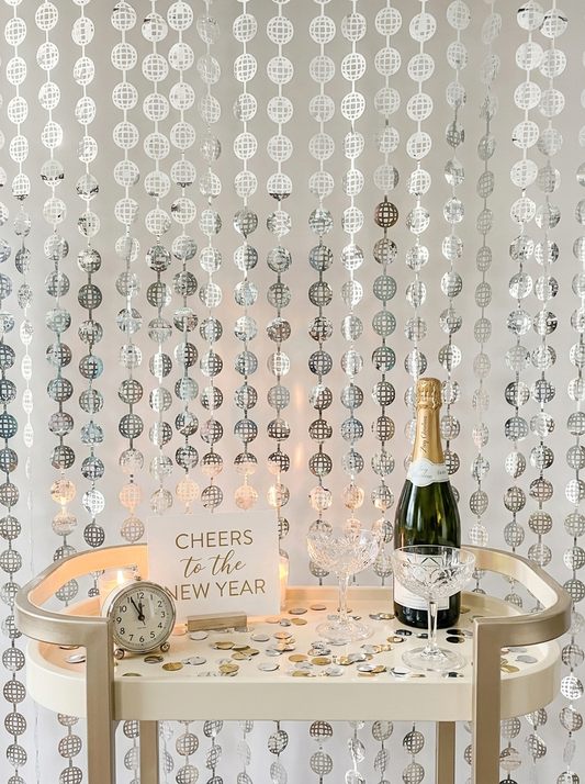 A festive New Year's Eve party setup featuring a champagne bottle, crystal glasses, a small clock, and a 'Cheers to the New Year' sign arranged on a cream-colored serving cart in front of a silver hanging beaded curtain backdrop.