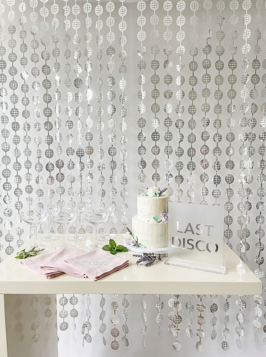 A silver beaded hanging party backdrop curtain displayed behind a white table decorated with a two-tiered cake, pink napkins, green leaves, and a white sign that reads 'LAST DISCO'.