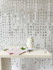 A silver beaded hanging party backdrop curtain displayed behind a white table decorated with a two-tiered cake, pink napkins, green leaves, and a white sign that reads 'LAST DISCO'.